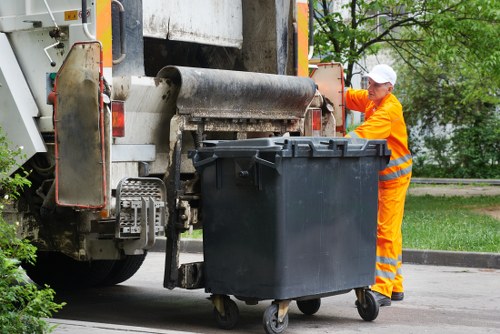 A waste collection operator wearing bright orange high-visibility clothing and a white safety helmet is positioned beside a large black plastic rubbish bin with a hinged lid and four wheels, which is situated on a paved street. The operator appears to be overseeing the collection process, possibly guiding the bin or preparing to move it. The refuse collection vehicle behind has a large, metallic compactor with visible textured rollers, and the rear section features hydraulic components and warning lights. Surrounding the scene, there are green leafy trees and a grassy area, suggesting a suburban or residential environment in St Albans, Hertfordshire. The lighting indicates a daylight setting, and the overall scene reflects typical rubbish collection operations carried out by companies like House Clearance St Albans in local neighbourhoods within the postal district postcodes associated with the town.