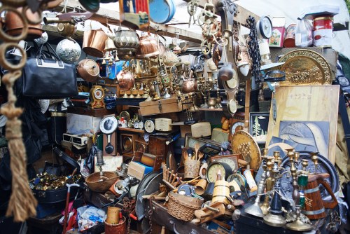 A densely packed outdoor display of assorted household items and decorative objects, including clocks, brass and copper cookware, small sculptures, lamps, and picture frames, arranged on shelves and tables. The objects vary in material, with many metal and wooden finishes, and are a mix of vintage and ornamental styles. The scene shows a cluttered marketplace or junk shop environment typical of secondhand goods or antique sales, with items overlapping and piled together. In the background, other merchandise is visible, including a framed picture or painting leaning against the display. The overall atmosphere is busy and eclectic, suggesting a location where household rubbish or unwanted items are collected or sold, consistent with services provided by companies like House Clearance St Albans in the Winchester postcode region. This detailed visual description aims to assist accessibility and support local SEO signals for rubbish removal service websites focusing on household clearance in the St Albans area.
