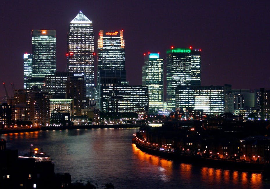 A nighttime cityscape of modern high-rise office buildings illuminated by numerous internal and external lights, situated along a river. The tallest skyscraper features a pointed, brightly lit roof st