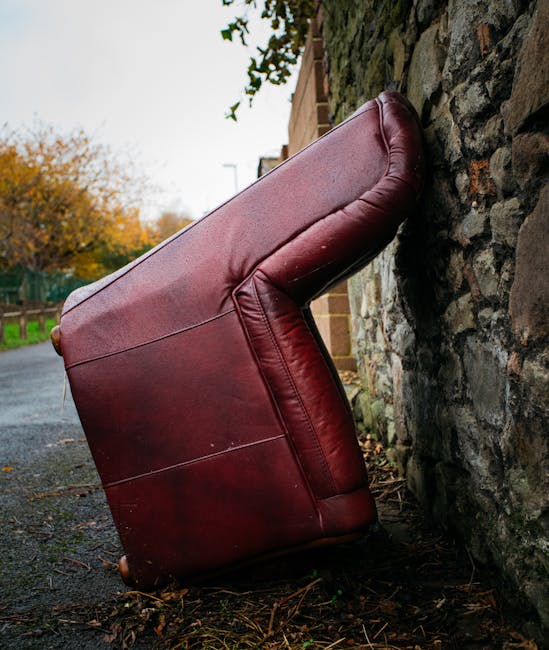 A detached, large red leather sofa is positioned upright and leaning against a rough stone wall outdoors, on a narrow pavement beside a residential street. The sofa's upholstery appears slightly worn 